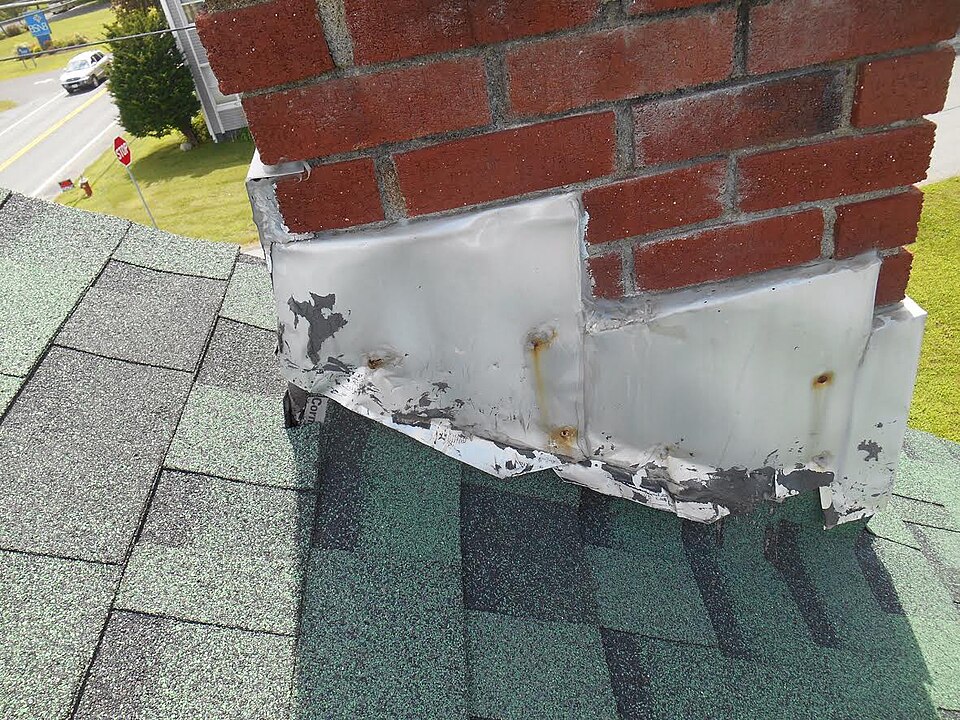 Close-up of a red brick chimney base where wrinkled, peeling white metal flashing is face-nailed into the brick and sealed with cracked gray caulk over asphalt shingles — an example of improper chimney flashing that invites roof leaks.